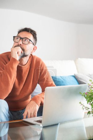 man sitting on sofa in front of an open laptop with thoughtful expression looking for a solution or idea. People and remote working lifestyle. Small business. Security password lost problems.