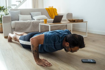 mature man working on push up, using phone and fitness online at home, copy space. Handsome middle-aged man in sportswear exercising in living room, attending personal trainer web class