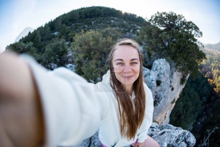 Foto de Young woman with blond hair taking a selfie portrait while hiking in the mountains - Happy hiker on top of a cliff smiling at camera - Travel and hobby concept - Imagen libre de derechos