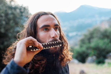 Photo for The musician enjoys playing the harmonica while standing on the street. a young man with long hair plays the harmonica. - Royalty Free Image
