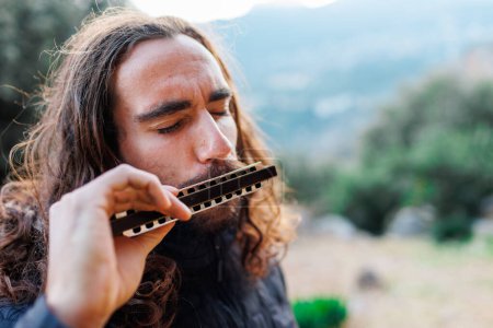 Photo for A young man with long hair plays the harmonica. a man enjoys playing the harmonica while standing on the street. - Royalty Free Image