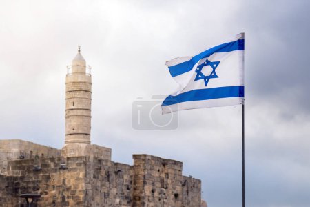 Foto de Bandera de Israel ondea en el viento con un minarete musulmán de una mezquita en el fondo en Jerusalén en el cielo nublado - Imagen libre de derechos