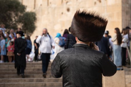 Foto de Vista trasera de un judío ortodoxo Hasidim con un gran sombrero de piel y un traje negro hablando por teléfono móvil cerca de la Cueva de los Patriarcas en la Ciudad Vieja de Hebrón en Cisjordania. - Imagen libre de derechos