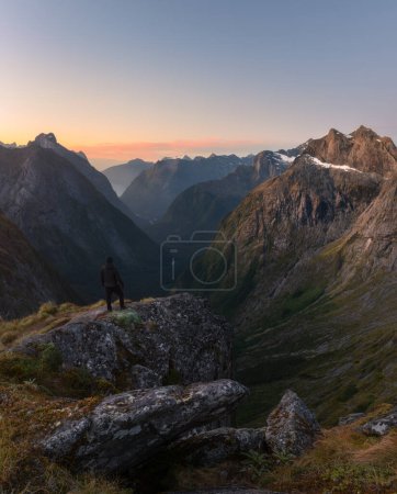 Young man standing on edge cliff facing to beautiful deep valley with rocky peaks at sunset