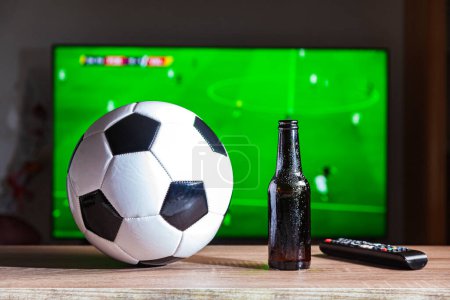 Photo for On a table is a soccer ball, a bottle of beer and a remote control. In the background, out of focus, there is a television set on which a football game is being broadcast. - Royalty Free Image
