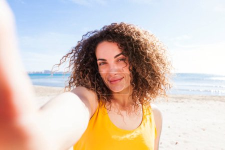 Beautiful young caucasian woman with curly hair taking a self portrait on the beach y a sunny day.