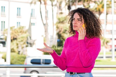 Photo for Angry young caucasian woman with curly hair is speaking with her smartphone on the street. - Royalty Free Image