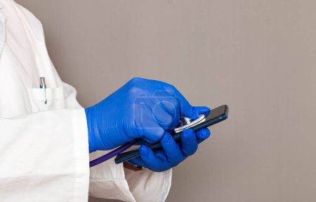 Foto de Primer plano de las manos de un médico irreconocible con guantes azules sosteniendo un teléfono inteligente mientras lo ausculta con un estetoscopio. - Imagen libre de derechos