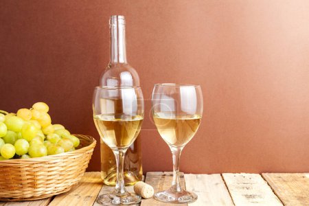 Téléchargez les photos : Nature morte d'un deux verres et une bouteille de vin blanc à côté d'un panier avec des raisins sur une table en bois rustique, fond brun. - en image libre de droit
