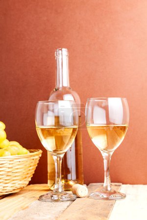 Téléchargez les photos : Pousse verticale de deux verres et une bouteille de vin blanc à côté d'un panier avec des raisins sur une table en bois rustique, fond brun. - en image libre de droit