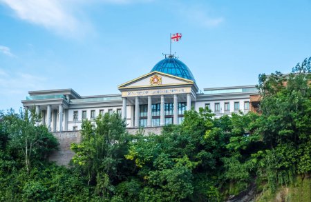 Foto de Presidential Palace in Tbilisi, Georgia with waving Georgian flag behind green trees. Palace of State Ceremonies or Avlabari Residence - modern building with glass dome in old town of Tiflis. - Imagen libre de derechos