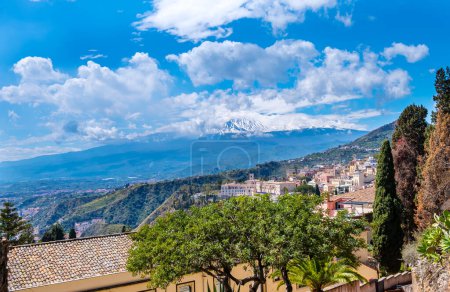 Taormina, Sizilien, Italien. Blick über die Stadt Taormina auf einem Hügel und den Vulkan Ätna zwischen Wolken am blauen Himmel. Beliebtes Touristenziel.