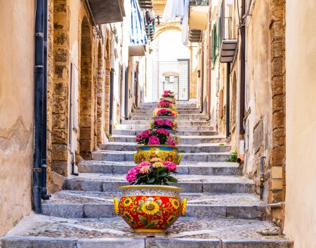 Photo for Narrow street in the old town of Cefalu, medieval village on Sicily island, Italy. Flower pots with traditional sicilian decoration. La Rocca cliff. Popular tourist attraction in Province of Palermo. - Royalty Free Image