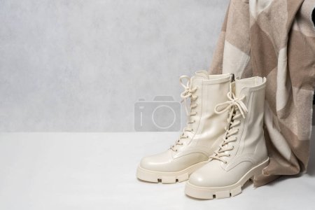 Photo for Pair of stylish white boots for women and beige winter scarf on light gray background. Trendy military boots on high platform with laces. Fall or winter female fashion and shoes still life. - Royalty Free Image