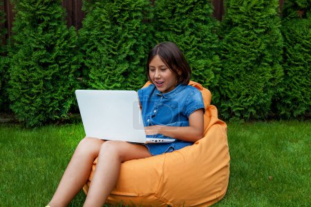 Das Mädchen sitzt auf einem Kissen mit offenem Laptop auf dem grünen Gras im Hof des Hauses. Mädchenhände auf der Tastatur. Fernlehrkonzept