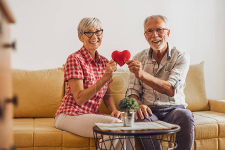 Photo for Senior couple sitting in living room and holding red heart shaped pillow. They are celebrating their anniversary. - Royalty Free Image