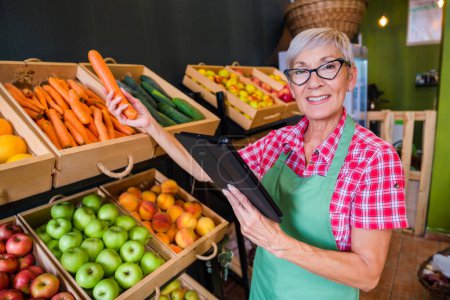 Photo for Mature woman is working at fruits and vegetables shop She is examining carrot. - Royalty Free Image