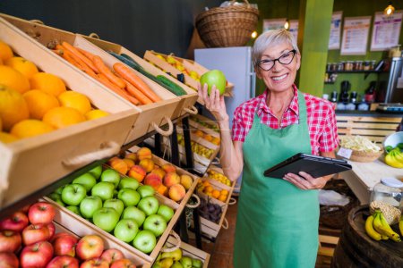 Photo for Mature woman is working at fruits and vegetables shop She is examining apples. - Royalty Free Image