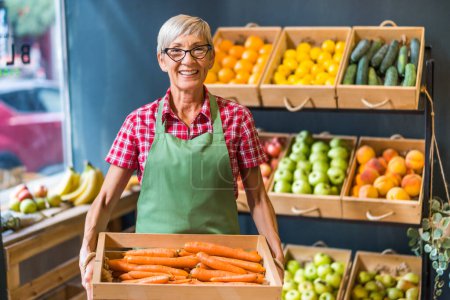 Photo for Mature woman works in fruits and vegetables shop. She is holding basket with carrot. - Royalty Free Image