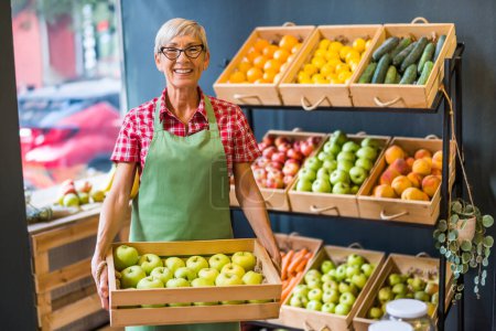 Mature woman works in fruits and vegetables shop. She is holding basket with apples.