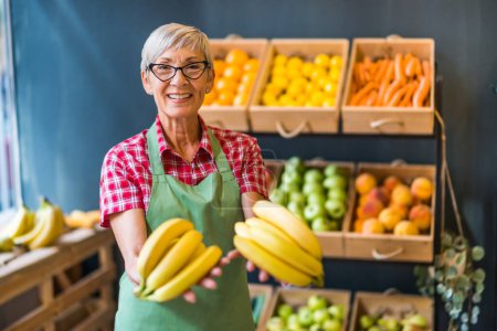 Photo for Mature woman works in fruits and vegetables shop. She is holding bananas. - Royalty Free Image