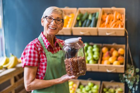 Photo for Woman works in fruits and vegetables shop. She is holding jar with peanuts. - Royalty Free Image