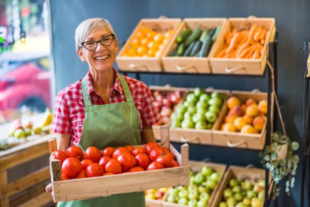Photo for Mature woman works in fruits and vegetables shop. She is holding basket with tomatoes. - Royalty Free Image