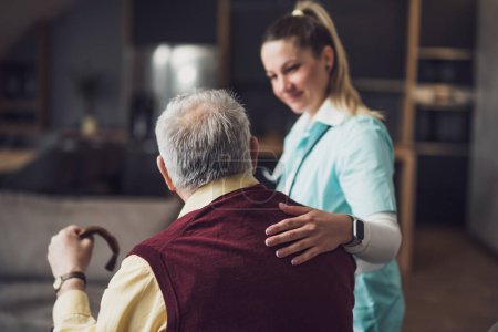 Téléchargez les photos : Le médecin à domicile rend visite à un aîné pour vérifier sa santé. Le soignant professionnel aide le vieil homme à son domicile. - en image libre de droit