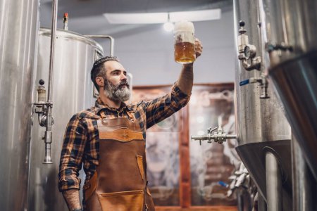 Maestro cervecero barbudo sosteniendo vaso de cerveza y evaluando sus características visuales. Pequeña empresa familiar, producción de cerveza artesanal.