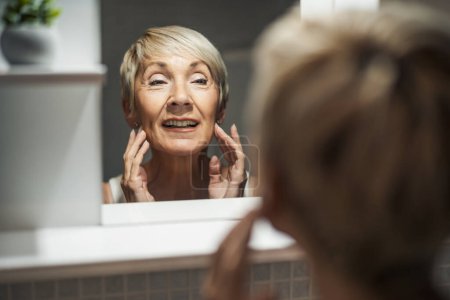 Téléchargez les photos : Femme mûre regardant son visage dans la salle de bain. Elle examine les rides de sa peau.. - en image libre de droit