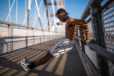 Junger afrikanisch-amerikanischer Mann übt auf der Brücke in der Stadt. Er macht umgekehrte Liegestütze.