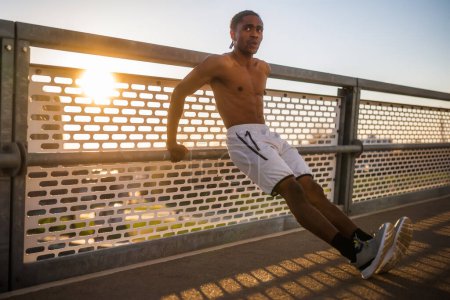 Un joven afroamericano se está ejercitando en el puente de la ciudad. Está haciendo flexiones inversas..