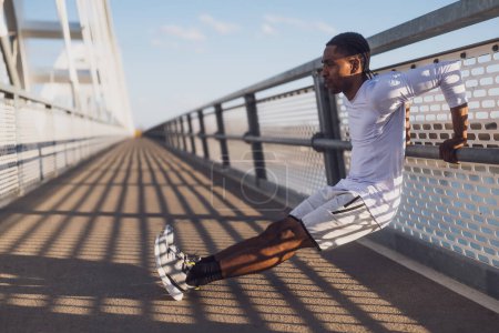 Un joven afroamericano se está ejercitando en el puente de la ciudad. Está haciendo flexiones inversas..