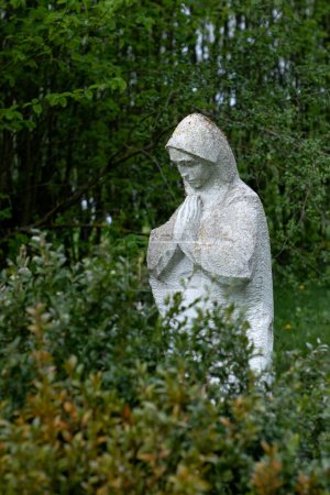 Una vieja estatua dañada de la Madre de Dios, abandonada en el cementerio arroja árboles borrosos con hojas verdes en el fondo.