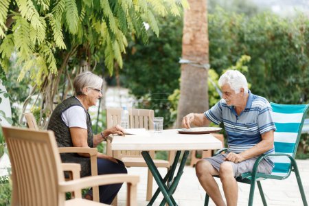 Téléchargez les photos : Aimer les conjoints âgés se reposant dans le jardin et profiter des jours, assis à l'extérieur.Livre de lecture femme, homme devant elle. - en image libre de droit