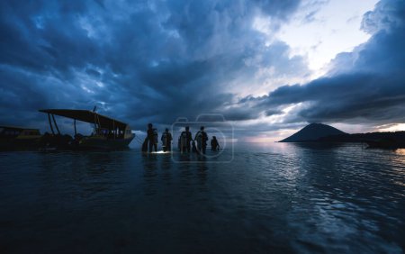 Téléchargez les photos : Plongeurs marchant dans l'océan avec des réservoirs et une torche pendant le coucher du soleil pour une plongée de nuit à Bunaken, Sulawesi, Indonésie - en image libre de droit