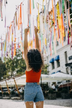 Photo for Happy young woman with curly brunette hair, standing on city streets, holding arms up, dancing. - Royalty Free Image