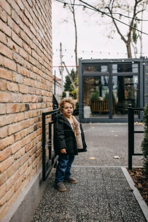 Foto de Pequeño niño feliz de pie en las calles de la ciudad, junto a una pared de ladrillo, en la entrada del jardín de infancia. - Imagen libre de derechos