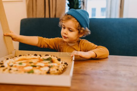 Photo for Little boy sitting at a cafe table opening a pizza box with traditional margherita pizza. - Royalty Free Image