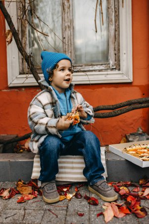 Téléchargez les photos : Petit garçon heureux mangeant de la pizza, à l'extérieur, assis dans la rue de la ville. - en image libre de droit