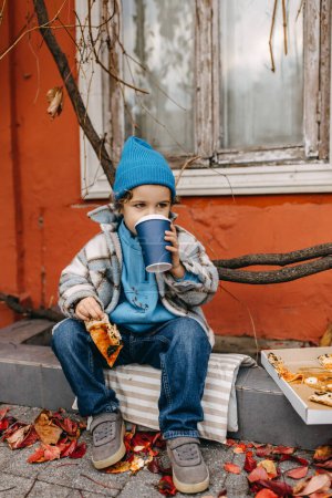 Téléchargez les photos : Petit garçon mangeant de la pizza et buvant du soda, dehors, assis dans la rue de la ville. - en image libre de droit