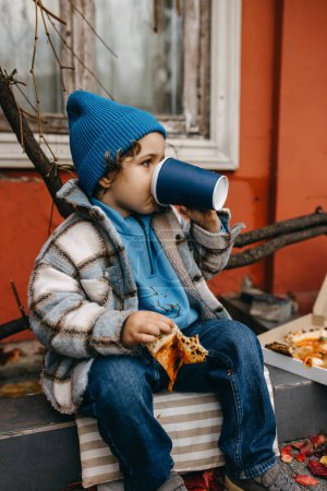 Téléchargez les photos : Little boy eating pizza and drinking soda, outdoors, sitting on city street. - en image libre de droit
