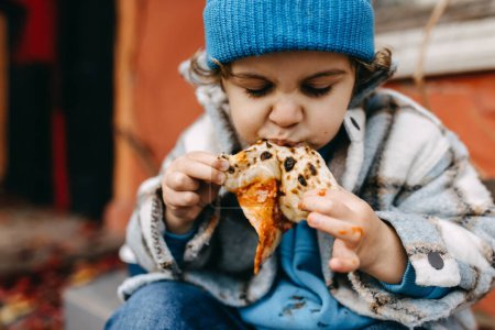 Photo for Closeup of a little boy eating a slice of pizza outdoors. - Royalty Free Image