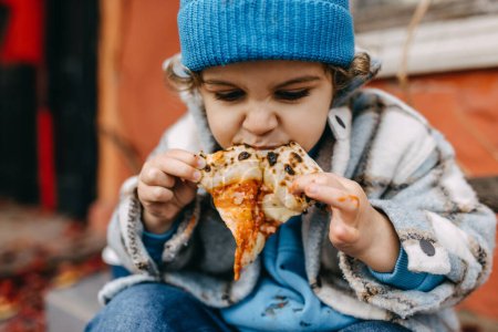 Photo for Closeup of a little boy eating a slice of pizza outdoors, with hands dirty in sauce. - Royalty Free Image