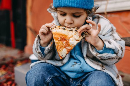 Téléchargez les photos : Closeup of a little boy eating a slice of pizza outdoors. - en image libre de droit