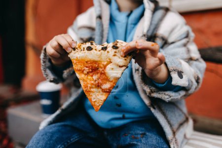 Foto de Closeup of a little boy holding a slice of pizza with dirty sauce hands, outdoors. - Imagen libre de derechos