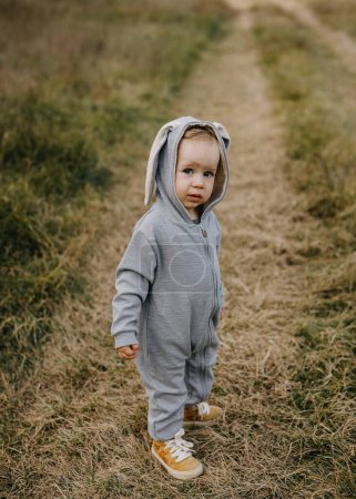 Téléchargez les photos : Petit enfant en costume de lapin gris debout sur l'herbe dans un champ ouvert, regardant la caméra. - en image libre de droit