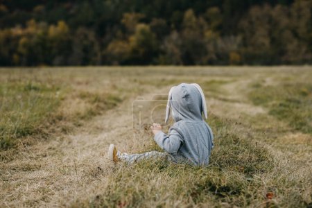 Téléchargez les photos : Petit enfant en costume de lapin gris assis sur l'herbe dans un champ ouvert. - en image libre de droit