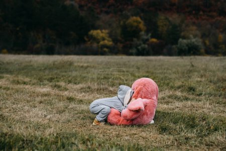 Foto de Niño pequeño con un disfraz de conejo, jugando con un gran juguete de conejo de felpa rosa, abrazándolo, al aire libre, en un campo abierto. - Imagen libre de derechos