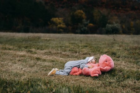 Foto de Little child wearing a bunny costume, laying with a big pink plush bunny toy, hugging it, outdoors, in an open field. - Imagen libre de derechos